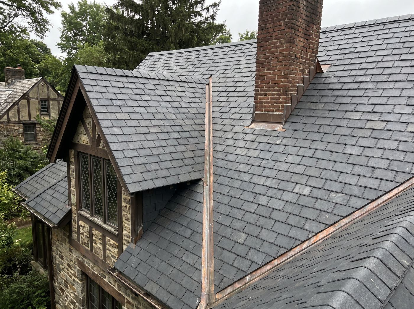 A steep period-revival roof with a dormer, valley flashing and a brick chimney