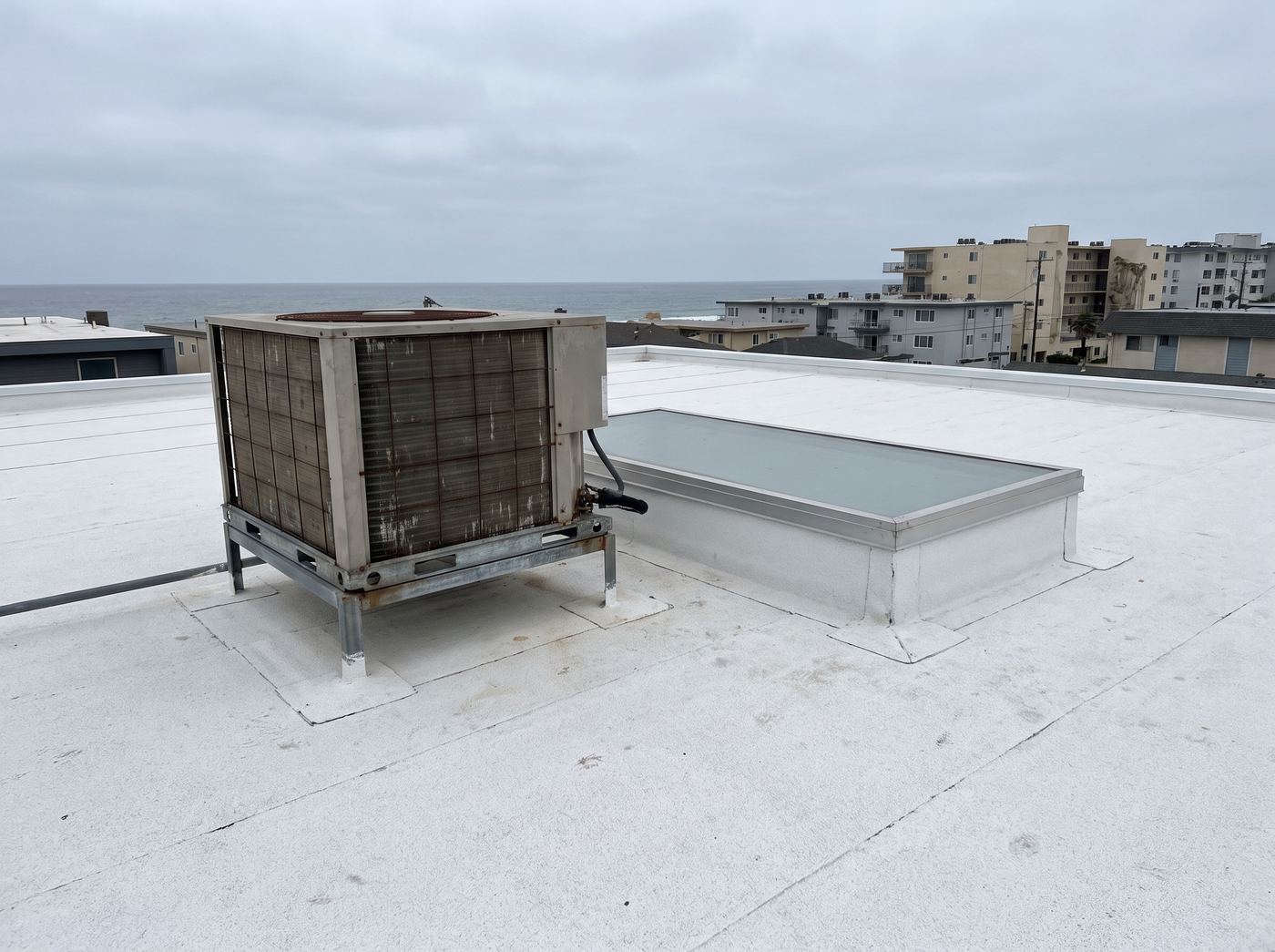 A flat roof deck near the ocean with a skylight curb and a rooftop unit on a stand