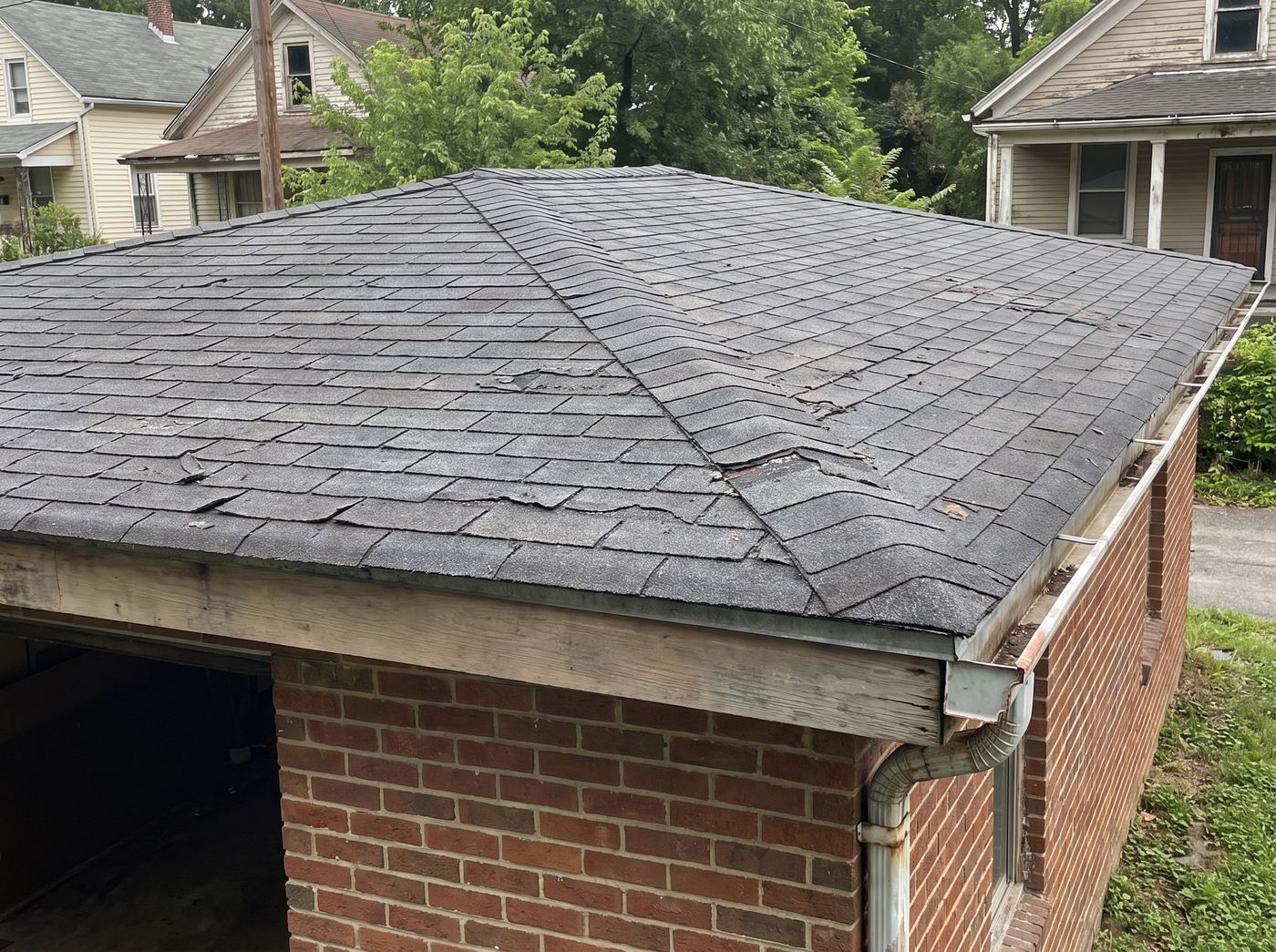 An aging composition shingle hip roof over a brick garage, with lifted and torn shingles along the hips and at the eave